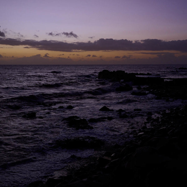 A light purple and yellow sunset, the wild ocean is in the foreground but is covered by the shadow 