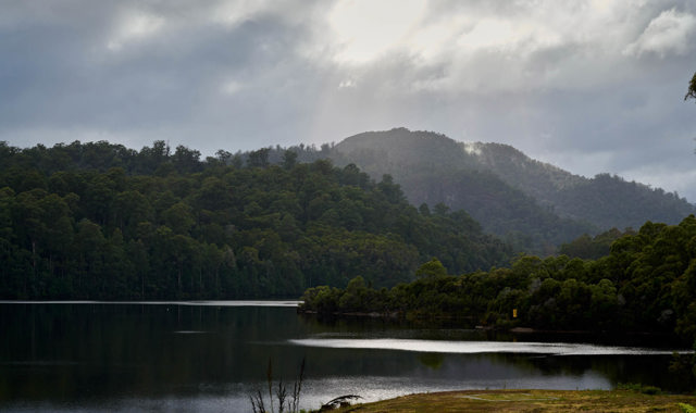 there is a glistening body of water, under beautiful sunlight, a wild mountainous backdrop and a yellow grass spot of land in the forground