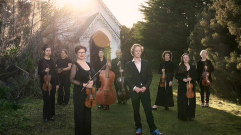 A group of people hold musical instruments standing on the grass in front of an old white small church, this church is surrounded by bushland