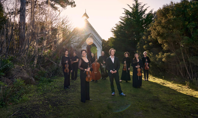 A group of people hold musical instruments standing on the grass in front of an old white small church, this church is surrounded by bushland