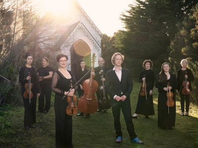 A group of people hold musical instruments standing on the grass in front of an old white small church, this church is surrounded by bushland